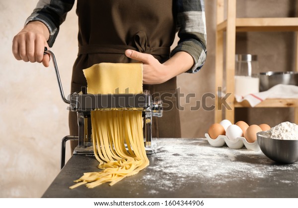 Woman preparing noodles with pasta maker machine\
at table indoors, closeup