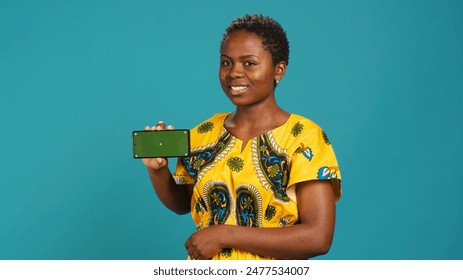Woman presenting her smartphone with isolated copy space screen, creating an advertisement while she wears african traditional attire with tribal elements. Gorgeous young adult in studio. Camera A. - Powered by Shutterstock