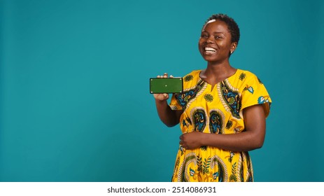 Woman presenting her smartphone with isolated copy space screen, creating an advertisement while she wears african traditional attire with tribal elements. Gorgeous young adult in studio. Camera B. - Powered by Shutterstock