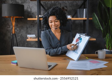 A woman is presenting a report on a laptop in front of a white wall. She is holding a piece of paper and pointing to it. The room has a lot of bookshelves and potted plants - Powered by Shutterstock