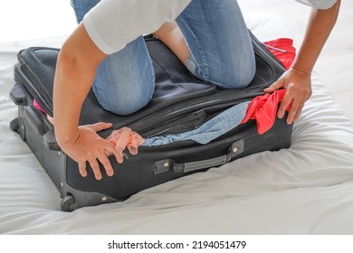 Woman Presses Her Foot On A Crowded Suitcase To Close It, Getting Ready For The Trip, Vacation, Travel. Woman Closes The Suitcase With Her Feet On Bed