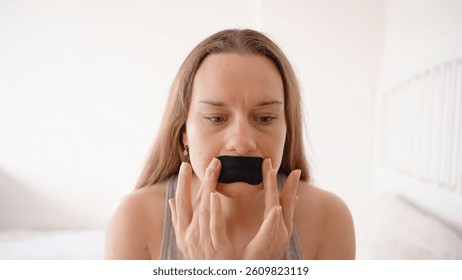 Mujer que presiona la cinta negra de la boca sobre los labios mientras que se sienta en la cama en la habitación blanca brillante, la aplicación diurna y el concepto de la respiración de la nariz