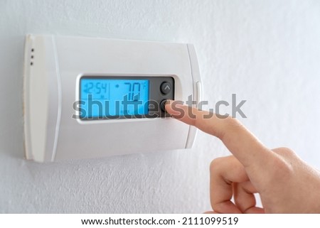 A woman is pressing the down button of a wall attached house thermostat with digital display showing temperature 70 degree Fahrenheit for heating, cooling, electricity and gas saving