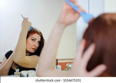 Woman In Process Of Hair Coloring At Home. Applying Red Color To The Hair And Looking In The Mirror. 