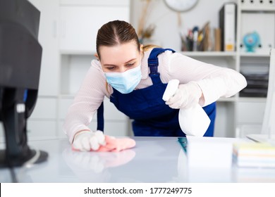 Woman professional cleaner in protective medical mask cleaning desk with detergents in office