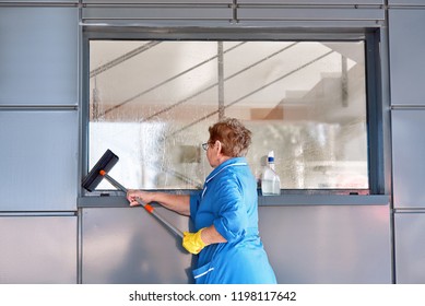 Woman From Professional Cleaning Service In Protective Rubber Yellow Gloves With Mop, Washing Large Window In Office Building. Cleaning Concept.  Employee Of Cleaning Company. Windows Clean