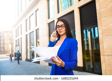 Woman Professional Government Worker In Classic Glasses And Suit Talking Via Mobile Phone And Reading Paper Documents While Standing Outdoors Against Modern Building Exterior 