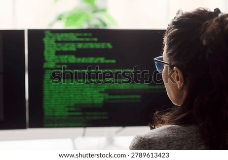 Woman programmer developer reading code on monitors. Back view in a tidy office, she analyzes syntax on a large screen, testing software and debugging code. Concept shows coding and focus.