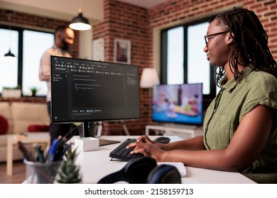 Woman programmer working remote writing code using computer while boyfriend is relaxing watching tv. African american freelancer developing software on pc sitting at desk in home living room