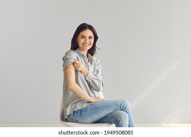 Woman promoting coronavirus vaccination and immunization campaign. Happy lady sitting on chair on gray studio background shows arm with adhesive patch after getting flu or Covid-19 vaccine injection