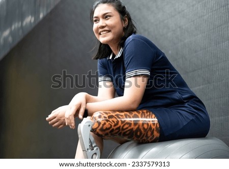 Woman with prosthetic leg sits isolated in gym. Asian female with foot prosthesis physical exercise in fitness. Artificial limb equipment help accident survivor or body injury people to mobility.