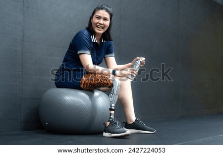 Woman with prosthetic leg sits relaxing on ball in gym. Asian female with foot prosthesis physical exercise in fitness. Mechanical artificial limb help orthopedic body injury people to strong mobility