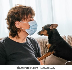 Woman Protected By A Mask Against Coronavirus, Covid-19 Or Any Other Disease, Sitting In Her Living Room With Her Surprised Dog Sniffing At Her