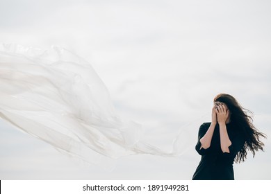 Woman protection. Female abuse. Humiliation violence. Portrait of insecure ashamed brunette lady in black hiding face with hands in white wind veil on cloudy fall sky art background with empty space.
