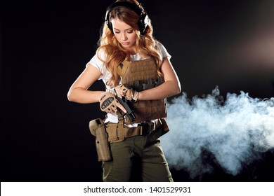 Woman in protective armour and headphones holds gun in hands, posing over dark smoky background. Army, dedication, weapon, technology.