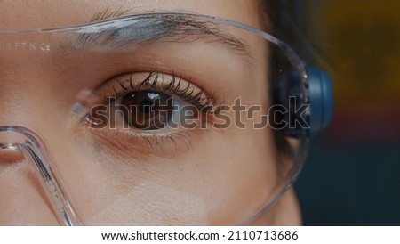Woman with protective glasses showing one eye in front of camera, brown eye blinking and focusing sight. Person wearing safety goggles, with half of face. Genetic anatomy. Close up