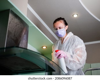 Woman in protective mask and glasses washes kitchen hood and furniture in the kitchen with disinfectant