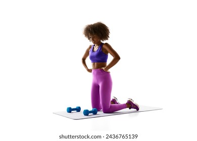 Woman in purple activewear kneeling on mat beside blue dumbbells, looking down against white studio background. Concept of sport, mourning routine, active and healthy lifestyle, energy, action.
