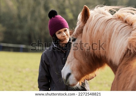 A woman in a purple bobble hat is making friends with a fawn-colored mare pony. The horse sniffs at the pocket of her gray yard jacket.
