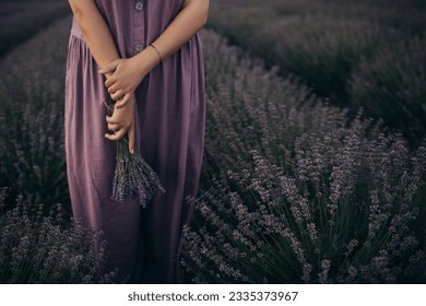 Woman in purple linen dress holding bunch of lavender flowers, standing in a lavender field at sunset. - Powered by Shutterstock