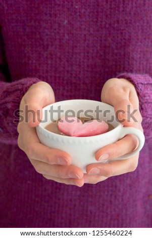 woman with purple pullover holding white cup filled with hot chocolate with a pink heart-shaped marshmallow