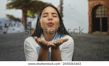 Woman purses lips in blowing pose within outdoor scene by a young latin hispanic figure, delivering kiss gestures filled with affection and crowned by a happy smile.