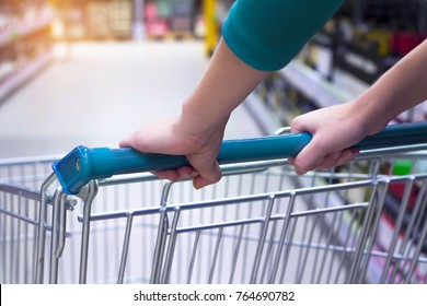 The Woman Pushes The Cart Along The Shop Aisle. Go With An Empty Red Basket With The Focus Of The Client's Focus. Hands Of The Girl On A Shopping Cart Close-up. Shopping At The Grocery Store