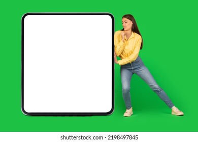 Woman Pushing Big Square Empty Gadget Screen Posing Standing Over Green Studio Background. Lady Trying To Move Large Mobile Phone Or Digital Tablet Display. Technology Concept. Mockup