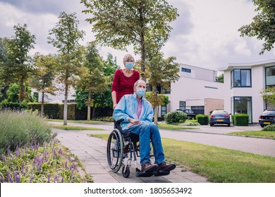 Woman Pushing Her Disabled Husband In His Wheelchair While On A Walk In The Neighborhood Wearing A Mask COVID-19/Corona.