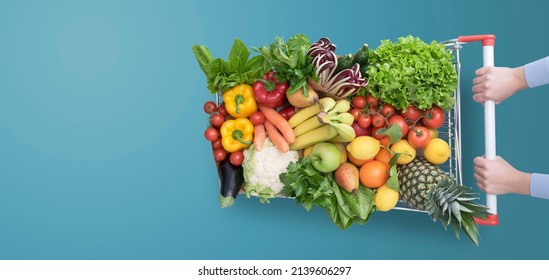 Woman pushing a shopping cart full of fresh delicious vegetables and fruits, grocery shopping concept, blank copy space