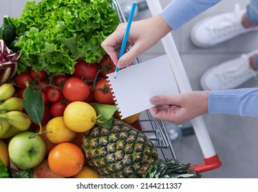 Woman pushing a shopping cart full of fresh greens and checking her shopping list, healthy food and grocery shopping concept - Powered by Shutterstock