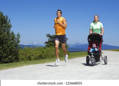 Woman Pushing A Stroller With Baby Walking Beside A Man Jogging, With Bushes And Trees In The Background