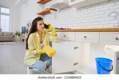 Woman Puts Bucket Pail Under Leaking Plastic Sink Pipe In Kitchen. Housewife Lady In Yellow Gloves Trying To Stop Leakage Problem, Holding Sponge And Calling Commercial Plumbing Service On Smartphone