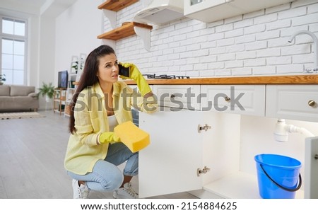 Woman puts bucket pail under leaking plastic sink pipe in kitchen. Housewife lady in yellow gloves trying to stop leakage problem, holding sponge and calling commercial plumbing service on smartphone