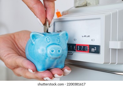 Woman Puts Coin In Piggy Bank Near A Gas Meter At Home. Natural Gas Consumption, Cost Of Utilities And Saving Concept