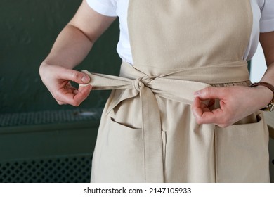 Woman puts on a linen beige apron over a white t-shirt. Deep green background - Powered by Shutterstock