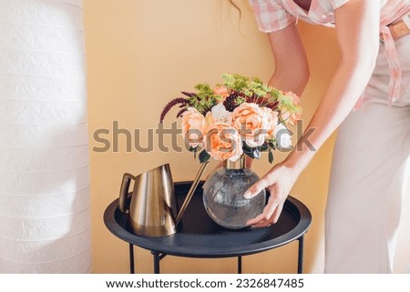Woman puts vase with bouquet of flowers on table at home. Floral arrangement with orange roses. Interior and summer decor