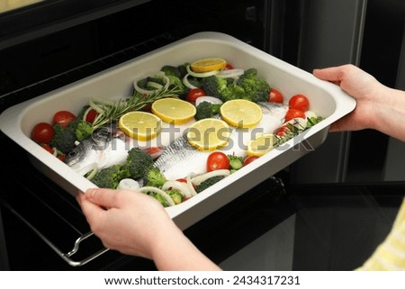 Woman putting baking dish with raw fish and vegetables into oven, closeup