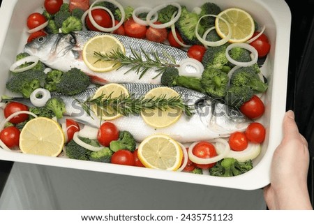 Woman putting baking dish with raw fish and vegetables into oven, top view