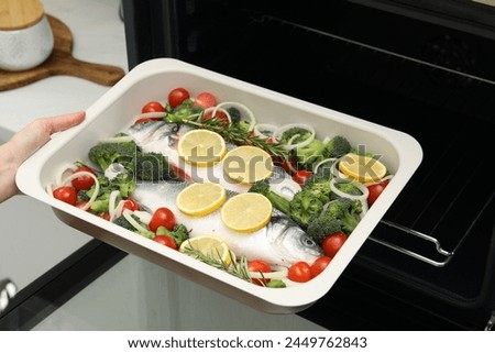 Woman putting baking dish with raw fish and vegetables into oven in kitchen, closeup