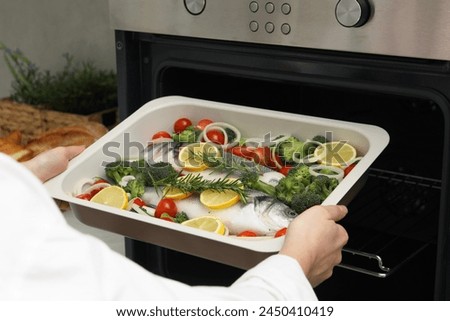 Woman putting baking dish with raw fish and vegetables into oven in kitchen, closeup