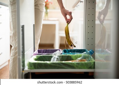 Woman Putting Banana Peel In Recycling Bio Bin In The Kitchen. Person In The House Kitchen Separating Waste. Different Trash Can With Colorful Garbage Bags.