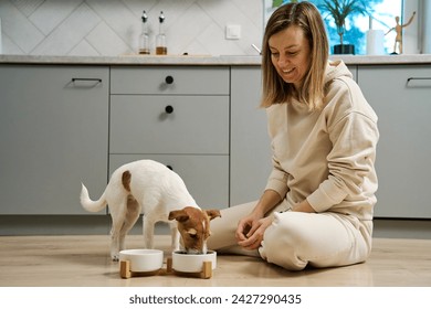 Woman putting food bowl with feed for her dog on the floor in kitchen, Female owner spending time together with pet at home, Animal feeding and pet care - Powered by Shutterstock