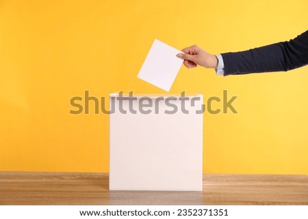 Woman putting her vote into ballot box on wooden table against orange background, closeup