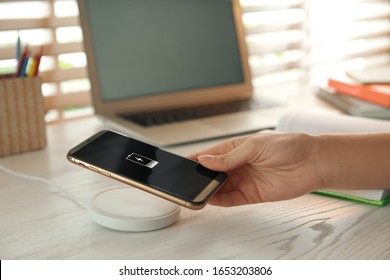 Woman putting mobile phone onto wireless charger at white wooden table, closeup. Modern workplace accessory