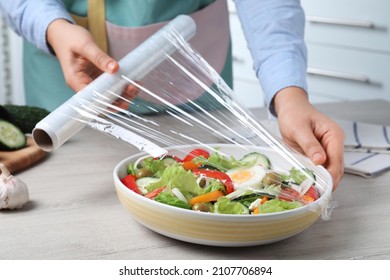 Woman putting plastic food wrap over bowl of fresh salad at wooden table indoors, closeup
