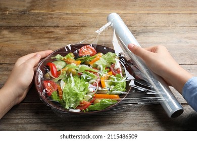 Woman Putting Plastic Food Wrap Over Bowl Of Fresh Salad At Wooden Table, Closeup