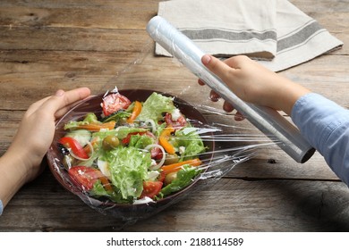 Woman Putting Plastic Food Wrap Over Bowl Of Fresh Salad At Wooden Table, Closeup