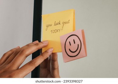 Woman putting a sticky note with phrase You Look Great Today! on the mirror. - Powered by Shutterstock