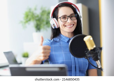 Woman radio host in headphones with microphone holds thumbs up. Recommendation of quality of work by streamer or presenter concept